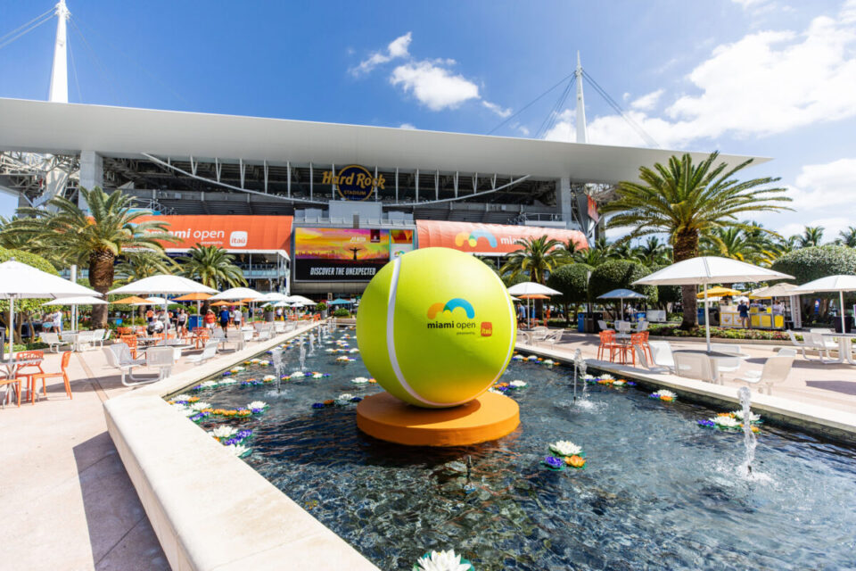 vibrant outdoor scene at the Miami Open outside Hard Rock Stadium, featuring a giant tennis ball sculpture with the event’s logo, a reflecting pool with floating flowers, palm trees, shaded seating areas, and event branding banners in the background under a bright blue sky.